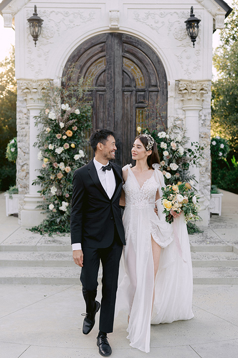  bride in a white lace gown with a flowing skirt with a high slit on the side, and tie straps.  The groom in a black michael kors tuxedo with a black bow tie 