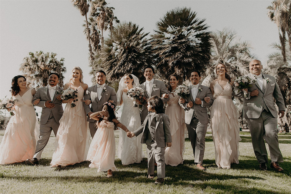  bride in a modern white gown with a v neckline and lace detailing, the groom in a light grey suit with a notch lapel and a white bow tie.  The groomsmen in light grey suits with blush bow ties and the bridesmaids in pink gowns 