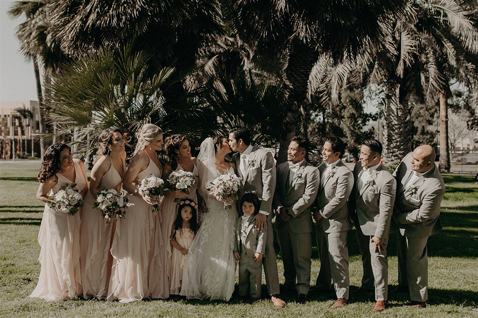  bride in a modern white gown with a v neckline and lace detailing, the groom in a light grey suit with a notch lapel and a white bow tie.  The groomsmen in light grey suits with blush bow ties and the bridesmaids in pink gowns 