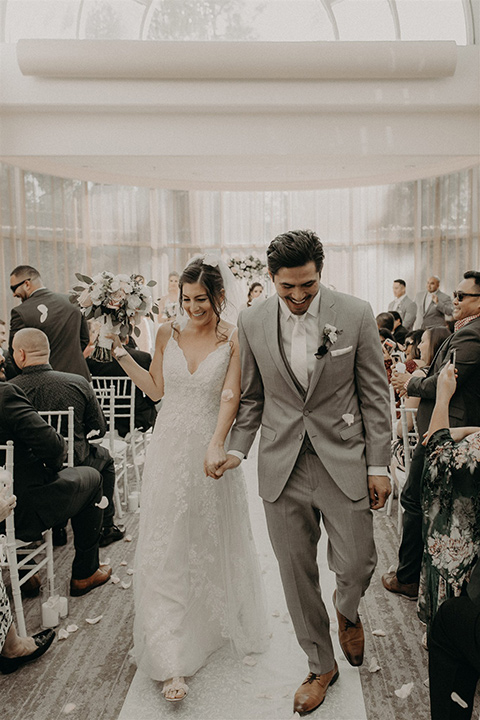  bride in a white lace gown with a v neckline and an elbow length veil and the groom in a light grey suit with a white long tie and brown shoes