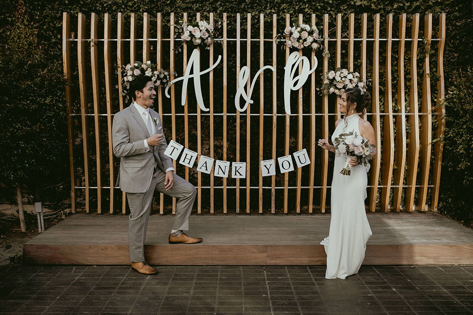  bride in a modern white gown with a high neckline, the groom in a light grey suit with a notch lapel and a white bow tie holding a welcome sign at the reception