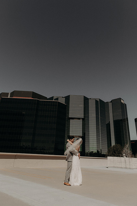  bride in a white lace gown with a v neckline and an elbow length veil and the groom in a light grey suit with a white long tie and brown shoes standing on the roof and kissing