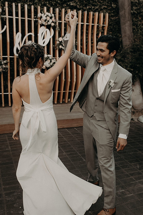  bride in a white lace gown with a v neckline and an elbow length veil and the groom in a light grey suit with a white long tie and brown shoes twirling