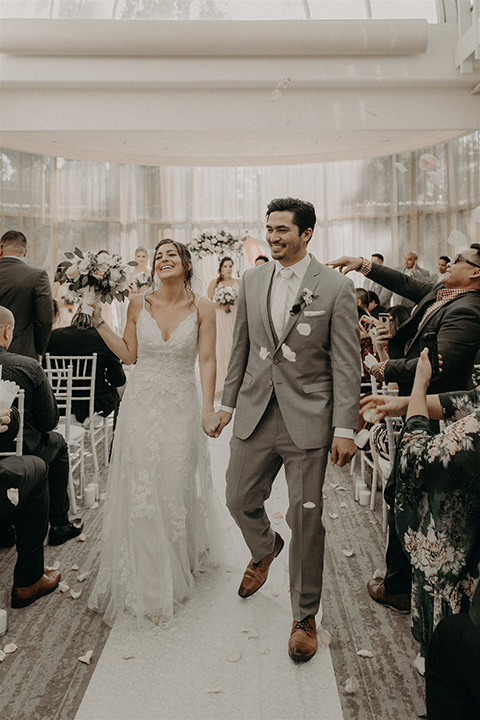  bride in a white lace gown with a v neckline and an elbow length veil and the groom in a light grey suit with a white long tie and brown shoes 