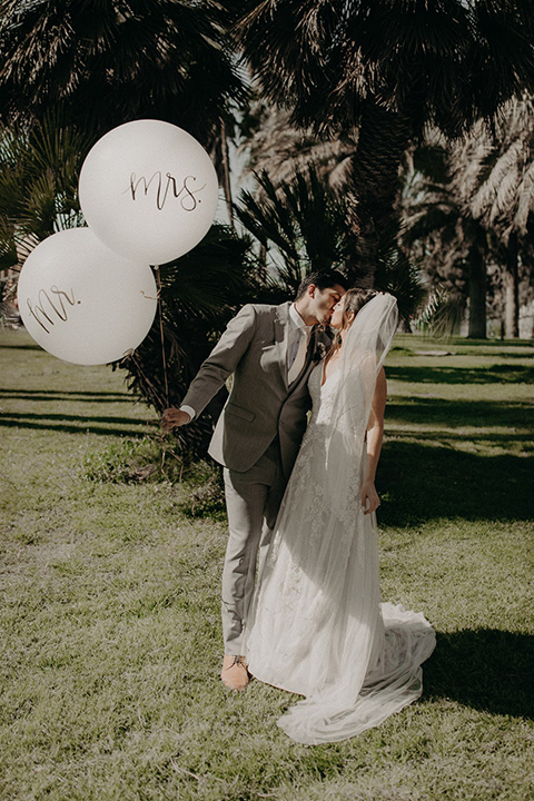  bride in a white lace gown with a v neckline and an elbow length veil and the groom in a light grey suit with a white long tie and brown shoes holding big balloons