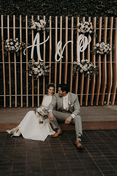  bride in a white lace gown with a v neckline and an elbow length veil and the groom in a light grey suit with a white long tie and brown shoes sitting on the ground