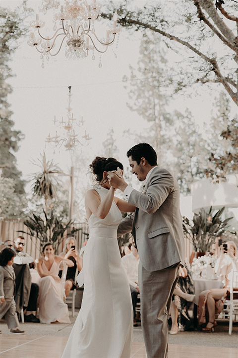  bride in a white modern gown with a high neckline and the groom in a light grey suit with a white long tie and brown shoes dancing