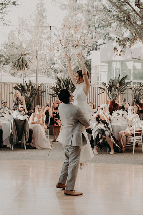  bride in a white modern gown with a high neckline and the groom in a light grey suit with a white long tie and brown shoes dancing 