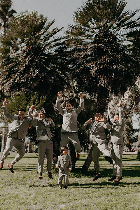  the groom in a light grey suit with a white long tie and brown shoes and groomsmen in a light grey suit with pink bow ties