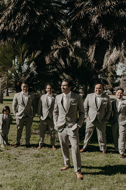  the groom in a light grey suit with a white long tie and brown shoes and groomsmen in light grey suits with pink bow ties