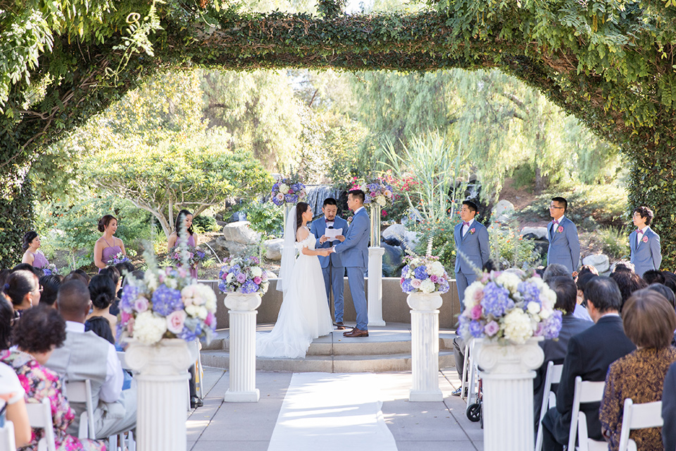  bride in a white gown with an off the shoulder neckline and the groom in a light blue suit with a blue bow tie, the groomsmen in light blue suits and the bridesmaids in lilac colored gowns 