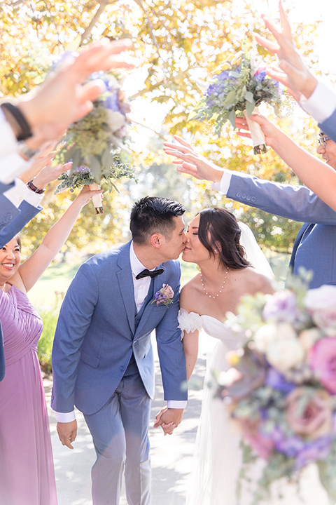  bride in a white gown with an off the shoulder neckline and the groom in a light blue suit with a blue bow tie, the groomsmen in light blue suits and the bridesmaids in lilac colored gowns