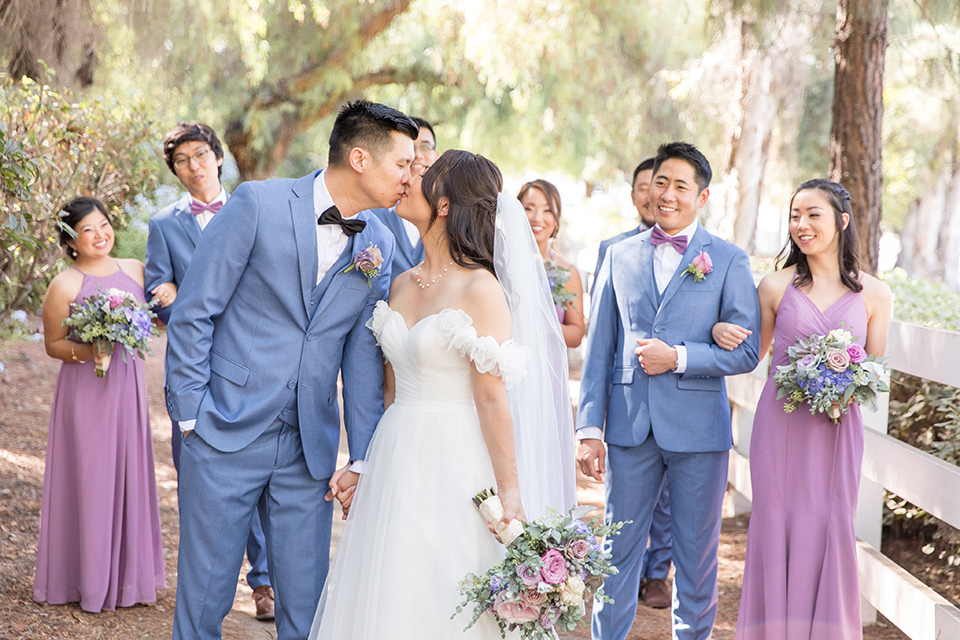  bride in a white gown with an off the shoulder neckline and the groom in a light blue suit with a blue bow tie, the groomsmen in light blue suits and the bridesmaids in lilac colored gowns 