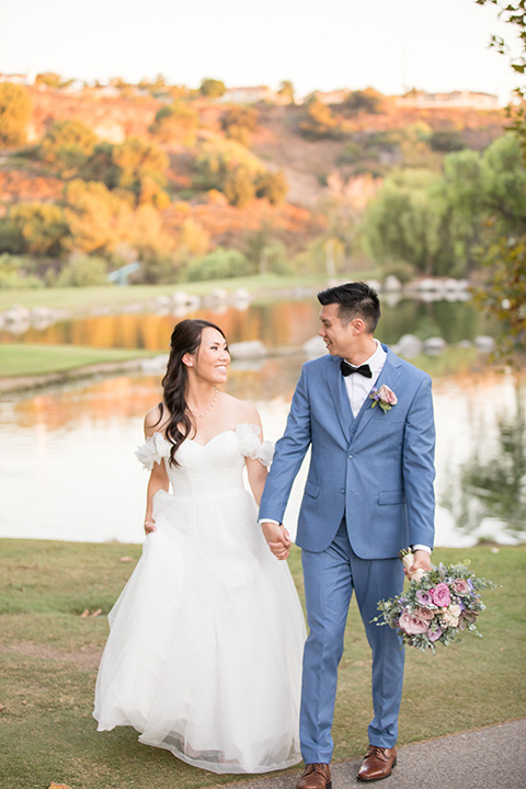  bride in a white gown with an off the shoulder neckline and the groom in a light blue suit with a blue bow tie, the groomsmen in light blue suits and the bridesmaids in lilac colored gowns 