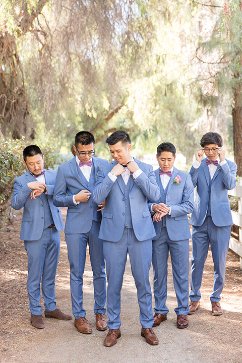  bride in a white gown with an off the shoulder neckline and the groom in a light blue suit with a blue bow tie, the groomsmen in light blue suits and the bridesmaids in lilac colored gowns 