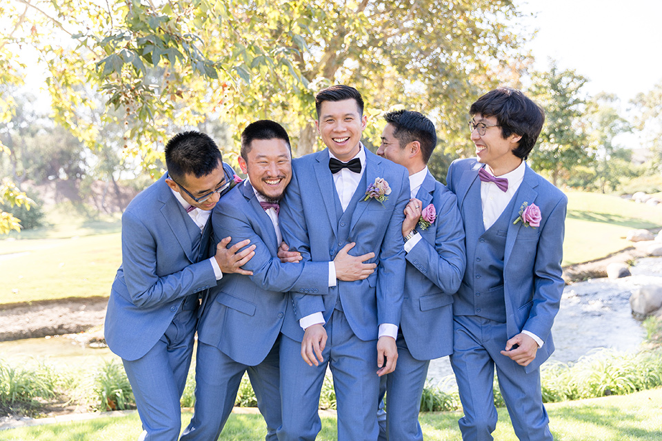  bride in a white gown with an off the shoulder neckline and the groom in a light blue suit with a blue bow tie, the groomsmen in light blue suits and the bridesmaids in lilac colored gowns 