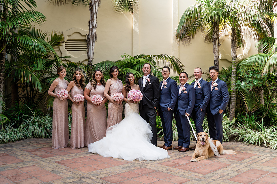  The bride in a mermaid style gown with a sweetheart neckline and the groom in a black tuxedo with a white long tie, the bridesmaids are in rose gold blush gowns and the groomsmen in navy blue suits with blush bow ties