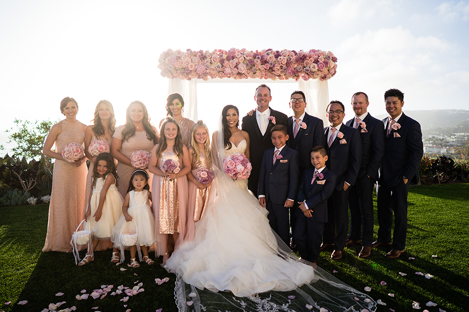  The bride in a mermaid style gown with a sweetheart neckline and the groom in a black tuxedo with a white long tie, the bridesmaids are in rose gold blush gowns and the groomsmen in navy blue suits with blush bow ties at the ceremony