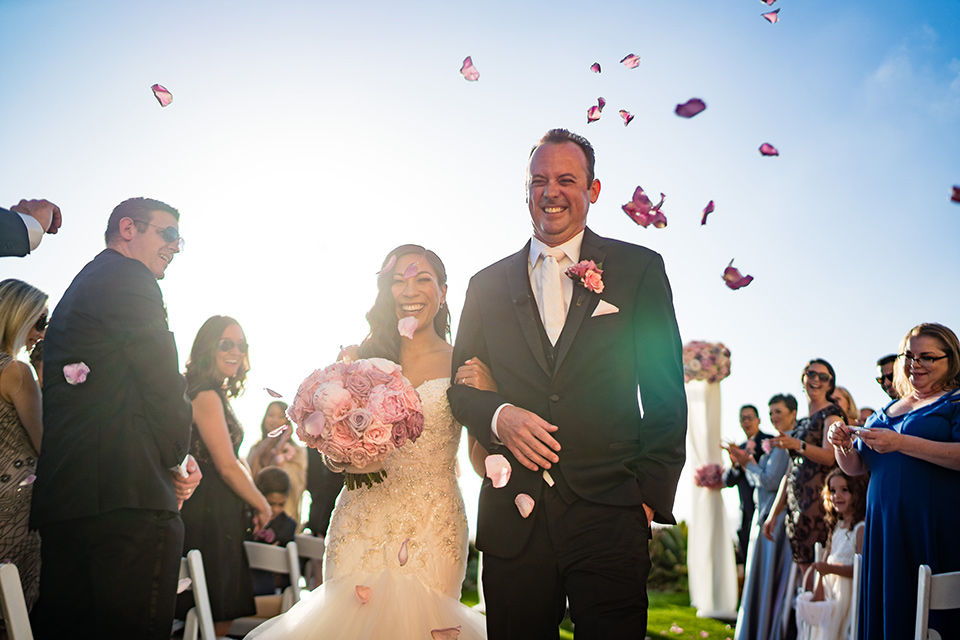  The bride in a mermaid style gown with a sweetheart neckline and the groom in a black tuxedo with a white long tie walking down the aisle 