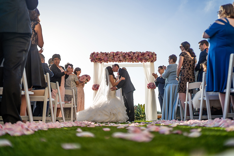  The bride in a mermaid style gown with a sweetheart neckline and the groom in a black tuxedo with a white long tie kissing at their ceremony 