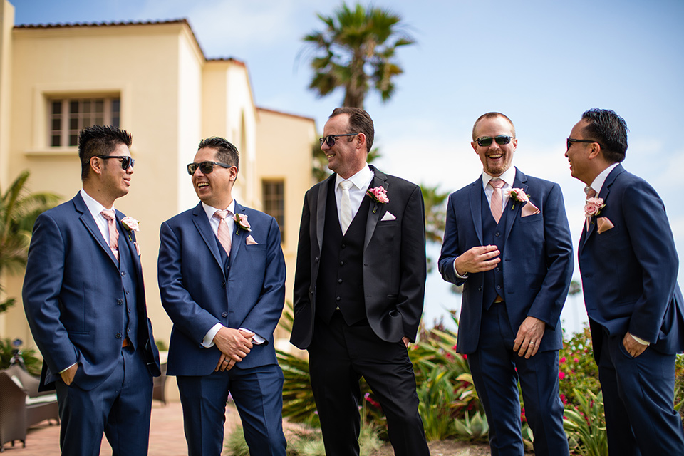  the groom in a black tuxedo with a white long tie and the groomsmen in navy blue suits with blush bow ties at the ceremony