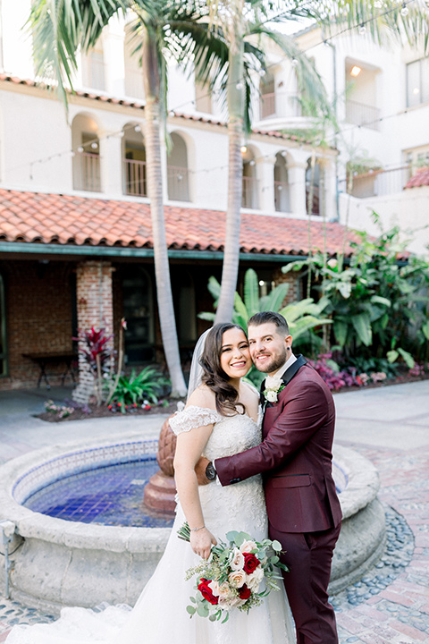  the bride in a lace a line gown with tulle cap sleeves and a modified sweetheart neckline, the groom in burgundy tuxedo with a shawl lapel tuxedo with a black long tie hugging by fountain