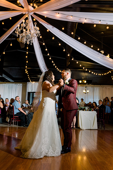  the bride in a lace a line gown with tulle cap sleeves and a modified sweetheart neckline, the groom in burgundy tuxedo with a shawl lapel tuxedo with a black long tie dancing the first dance