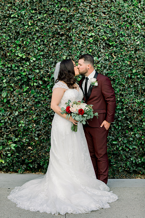  the bride in a lace a line gown with tulle cap sleeves and a modified sweetheart neckline, the groom in burgundy tuxedo with a shawl lapel tuxedo with a black long tie kissing by ivy wall