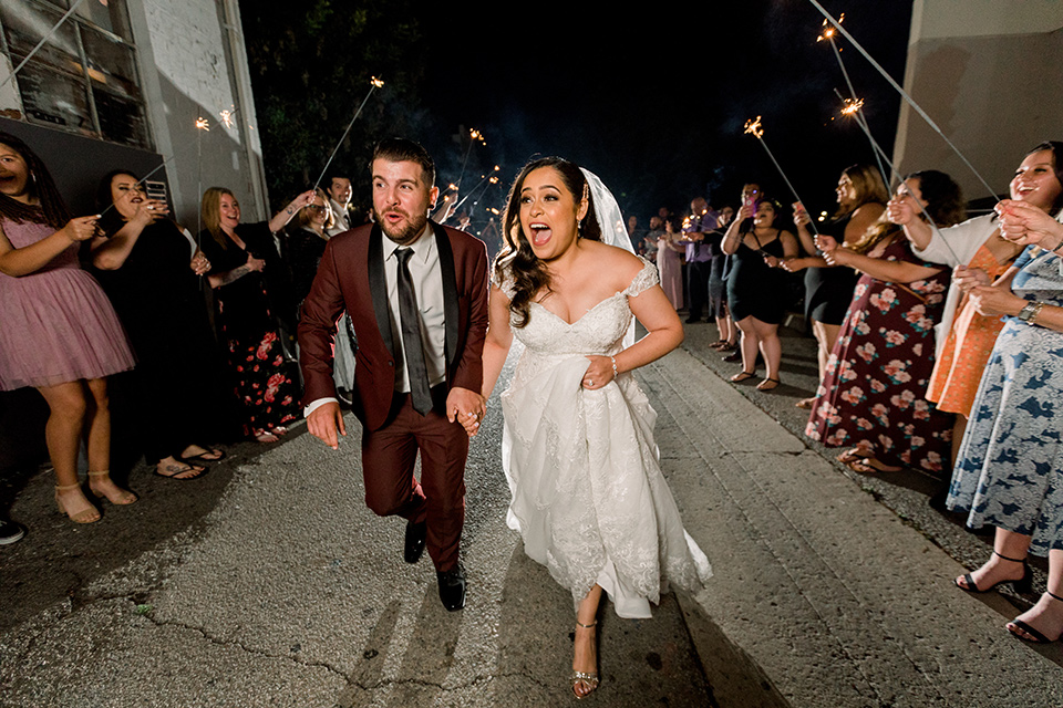  the bride in a lace a line gown with tulle cap sleeves and a modified sweetheart neckline, the groom in burgundy tuxedo with a shawl lapel tuxedo with a black long tie smiling and leaving the wedding with the guests holding sparklers