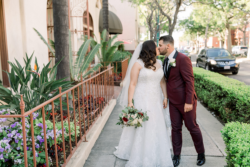  the bride in a lace a line gown with tulle cap sleeves and a modified sweetheart neckline, the groom in burgundy tuxedo with a shawl lapel tuxedo with a black long tie kissing outside the venue