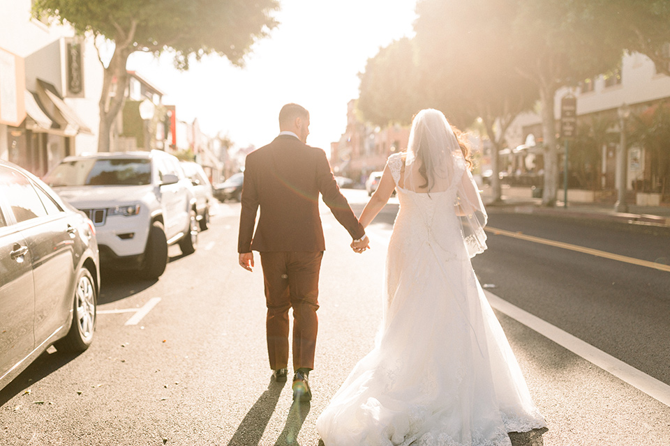  the bride in a lace a line gown with tulle cap sleeves and a modified sweetheart neckline, the groom in burgundy tuxedo with a shawl lapel tuxedo with a black long tie walking away from the camera down the street