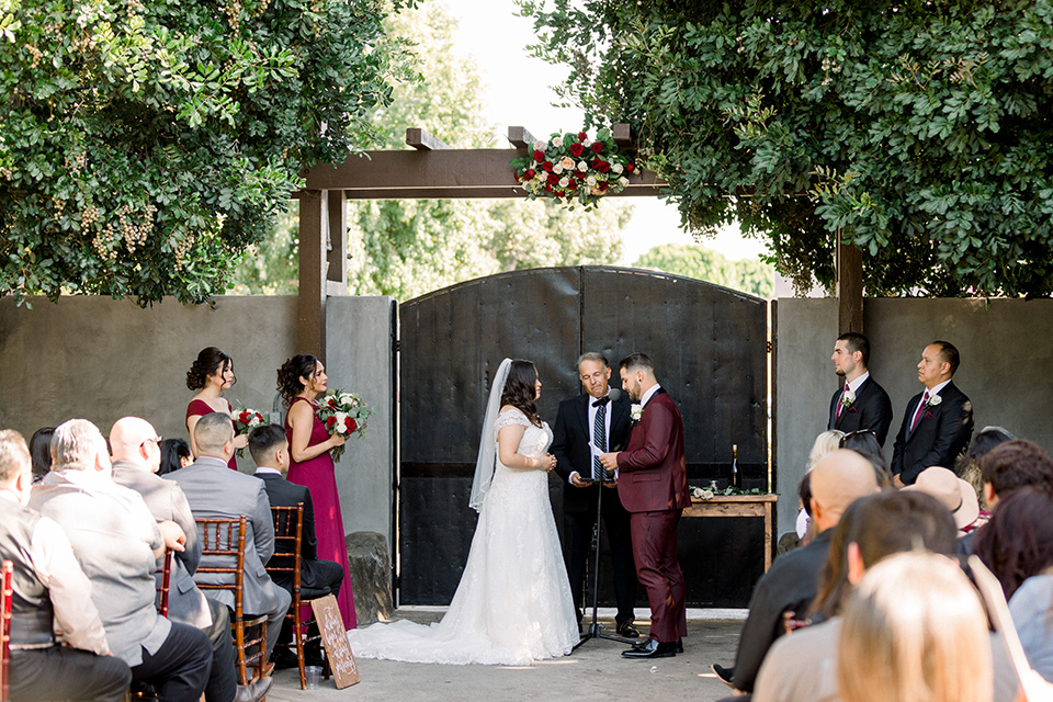  the bride in a lace a line gown with tulle cap sleeves and a modified sweetheart neckline, the groom in burgundy tuxedo with a shawl lapel tuxedo with a black long tie at the ceremony