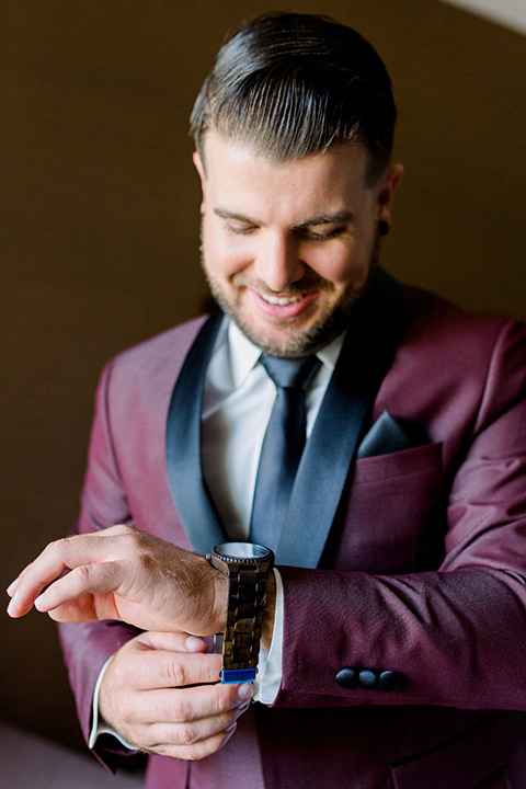  the groom in burgundy tuxedo with a shawl lapel tuxedo with a black long tie looking at watch