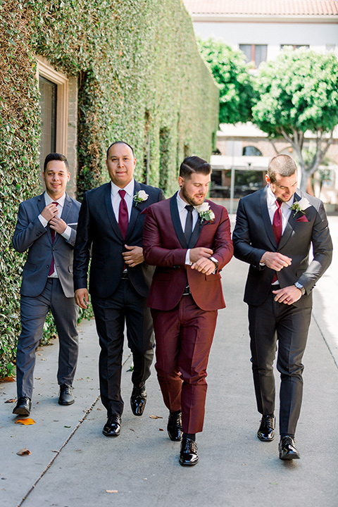  the groom in burgundy tuxedo with a shawl lapel tuxedo with a black long tie and groomsmen in black and grey suits with long burgundy ties walking towards camera