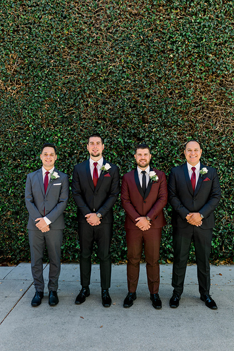  the groom in burgundy tuxedo with a shawl lapel tuxedo with a black long tie and his groomsmen in black and grey suits with burgundy long ties