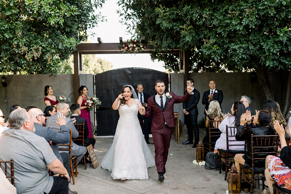  the bride in a lace a line gown with tulle cap sleeves and a modified sweetheart neckline, the groom in burgundy tuxedo with a shawl lapel tuxedo with a black long tie at the ceremony