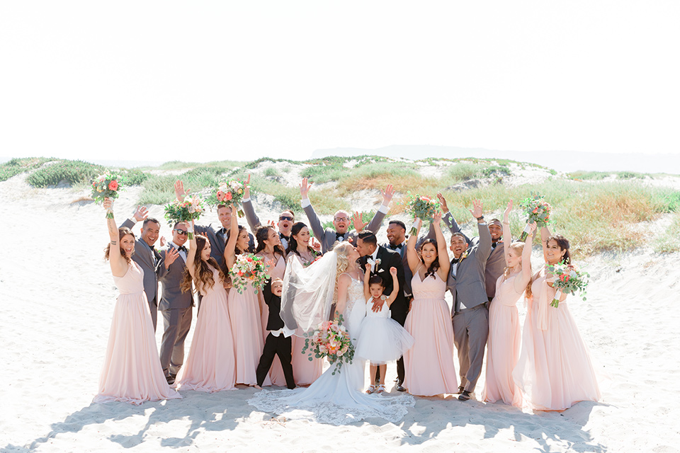  bride in a lace formfitting gown and a long veil, the groom in a black tuxedo with a black bow tie, the bridesmaids in blush pink gowns, and the groomsmen in light grey suits with black bow ties
