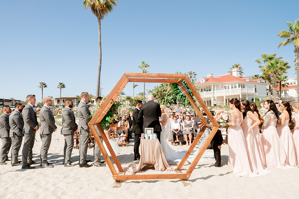  bride in a lace formfitting gown and a long veil, the groom in a black tuxedo with a black bow tie, the bridesmaids in blush pink gowns, and the groomsmen in light grey suits with black bow ties with the ceremony on the sand