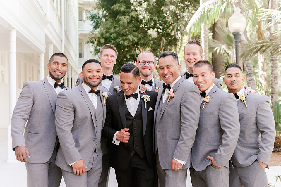  the groom in a black tuxedo with a black bow tie and the groomsmen in light grey suits with black bow ties laughing