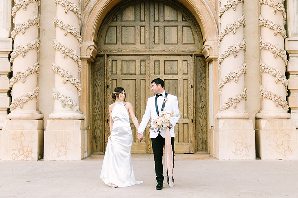  bride in a white gown with a high neckline and gold headpiece and the groom in a white shawl lapel tuxedo with a black bow tie 