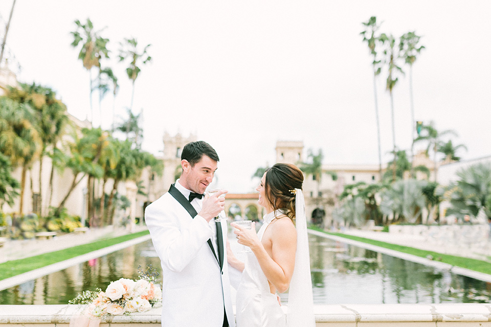  bride in a white gown with a high neckline and gold headpiece and the groom in a white shawl lapel tuxedo with a black bow tie 