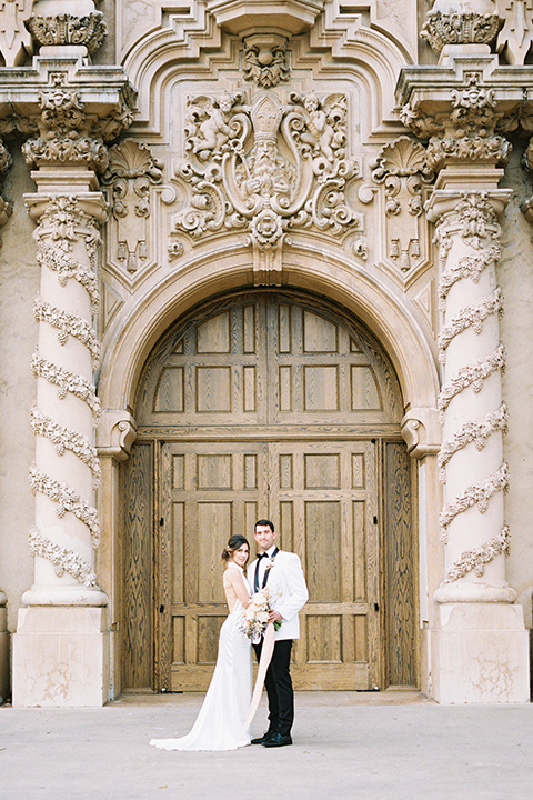  bride in a white gown with a high neckline and gold headpiece and the groom in a white shawl lapel tuxedo with a black bow tie