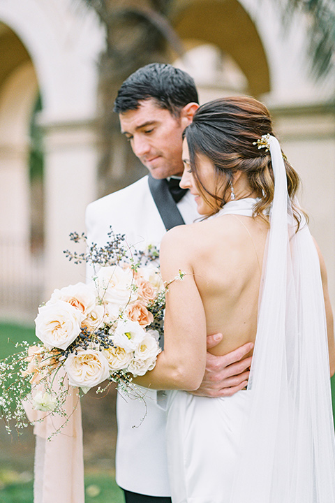  bride in a white gown with a high neckline and gold headpiece and the groom in a white shawl lapel tuxedo with a black bow tie