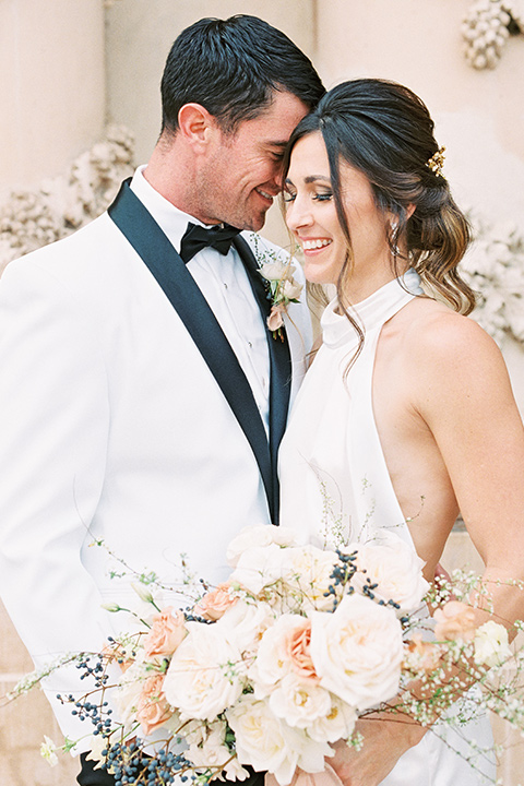  bride in a white gown with a high neckline and gold headpiece and the groom in a white shawl lapel tuxedo with a black bow tie