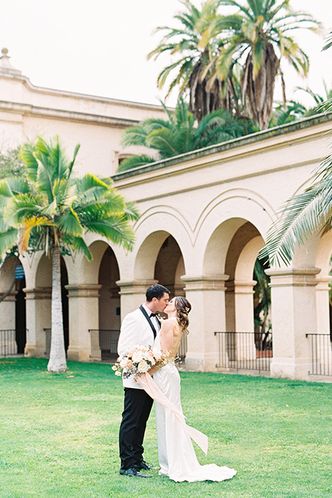  bride in a white gown with a high neckline and gold headpiece and the groom in a white shawl lapel tuxedo with a black bow tie