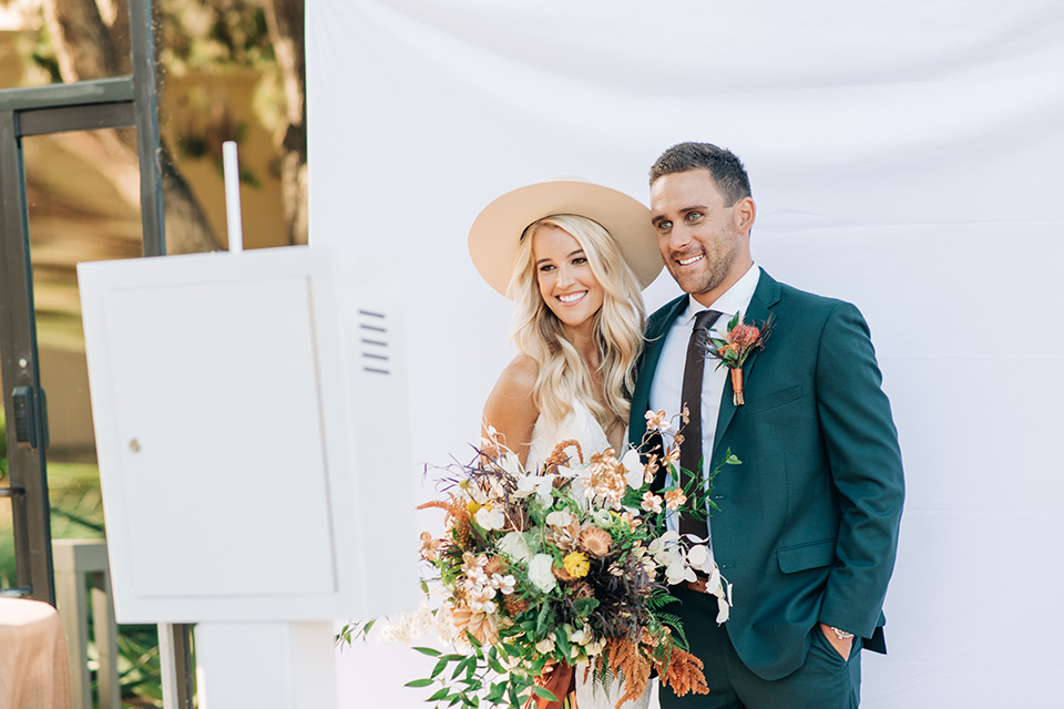 bride in a white lace bohemian style wedding gown with a tan wide brimmed hat and the groom in a dark green suit with a chocolate long tie