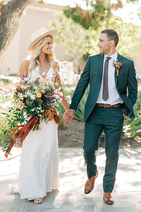  bride in a white lace bohemian style wedding gown with a tan wide brimmed hat and the groom in a dark green suit with a chocolate long tie 