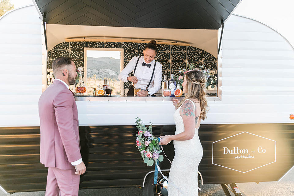  bride in a white lace bohemian style gown with a sweetheart neckline and thin straps and the groom in a rose pink suit with a burgundy bow tie at bar cart