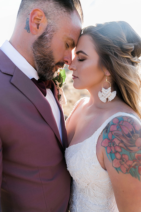  bride in a white lace bohemian style gown with a sweetheart neckline and thin straps and the groom in a rose pink suit with a burgundy bow tie