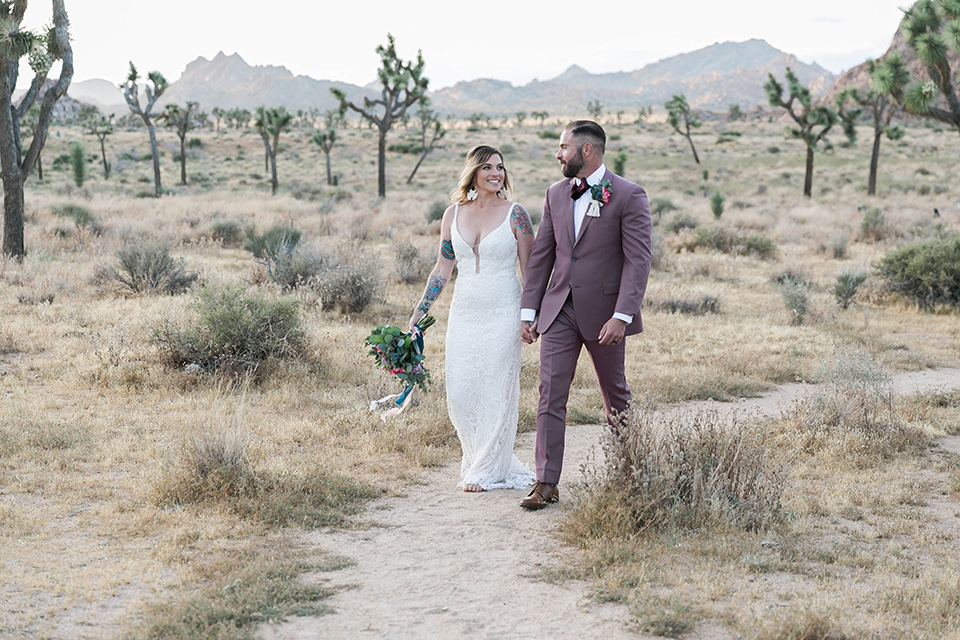  bride in a white lace bohemian style gown with a sweetheart neckline and thin straps and the groom in a rose pink suit with a burgundy bow tie walking together